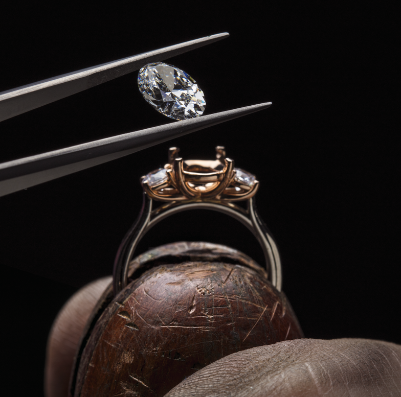 Jeweler holding a diamond with precision tweezers, preparing to set it into an intricate ring setting. This close-up highlights the artistry and craftsmanship involved in creating custom jewelry at First Person Jewelers.